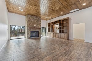 A living room featuring wood floors and celing, with a stone fireplace and built in shelving on the wall. There are two sliding doors on either side of the fireplace, leading to an outdoor patio.